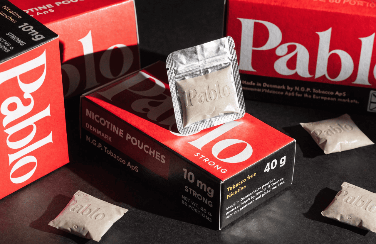A can of Pablo nicotine pouches in front of a factory with a Danish flag.